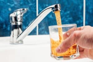 A hand holds a glass into which rusty dirty water is pouring from a tap.