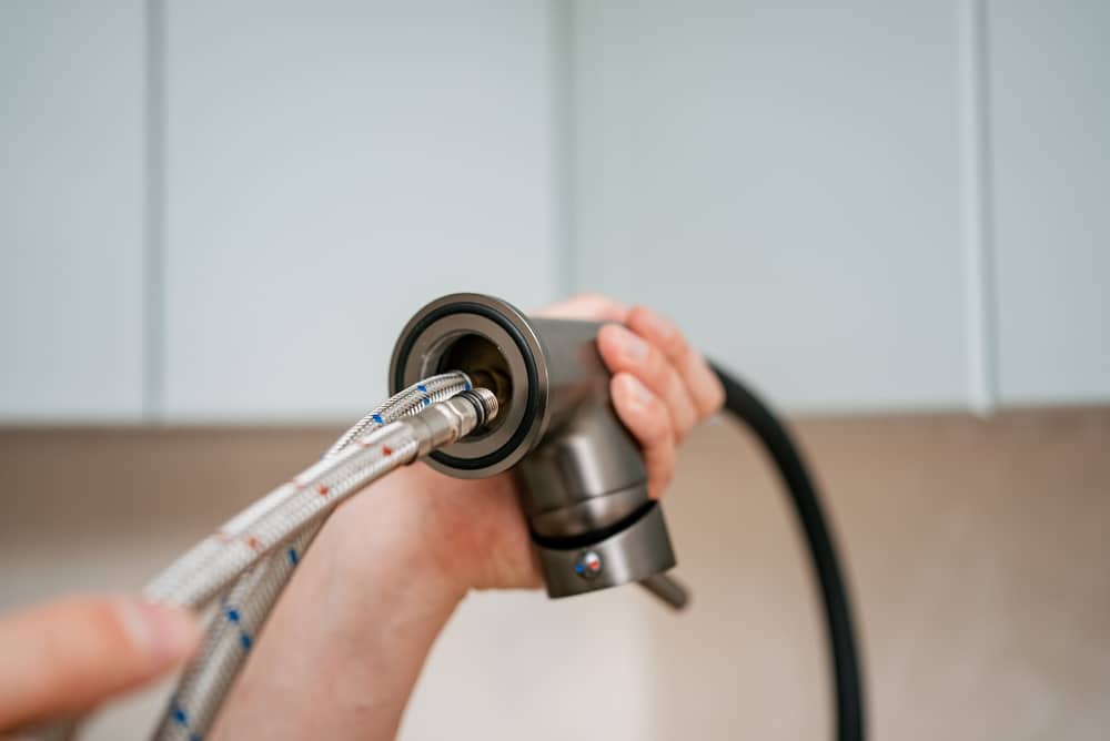 Close-up of a plumber's hands connecting flexible water supply lines to the base of a new kitchen faucet, demonstrating the process of plumbing installation in a modern kitchen environment