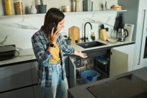 Caucasian young adult woman standing in modern kitchen talking on smartphone gesturing toward open cabinet under sink appearing to discuss plumbing issue or home maintenance
